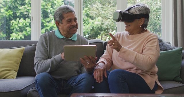 Elderly couple sitting on couch in living room enjoying modern technology. The elderly man is holding a tablet while the elderly woman is using a VR headset, displaying joy and engagement. Ideal for articles on technology adoption among seniors, digital inclusion, healthy aging with technology, or lifestyle of retired individuals.