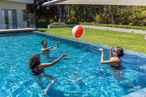Multicultural women playing with beach ball in modern backyard pool on sunny summer day