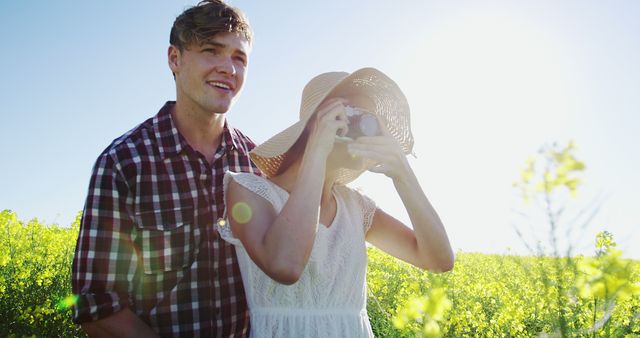 A young couple is enjoying a bright and sunny day together in a field. The woman is wearing a sunhat and using a vintage camera, capturing the moment, while the man, dressed in a plaid shirt, stands close by with a smile. This scene evokes feelings of warmth, carefree summer days, and youthful amusement. Perfect for use in advertisements for travel, lifestyle, outdoor activities, or romantic getaways.