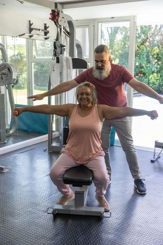 Senior woman performing seated lateral raises with trainer coaching in home gym