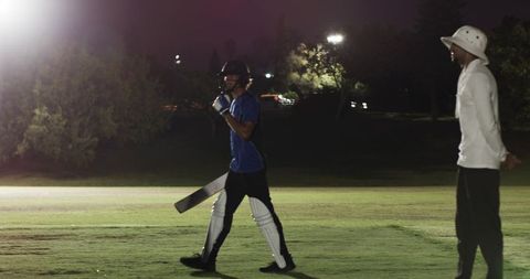 Cricketer walking off night pitch wearing helmet and pads under bright floodlights in match