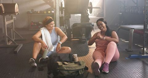 Diverse female friends laughing and tapping smartwatches after gym workout