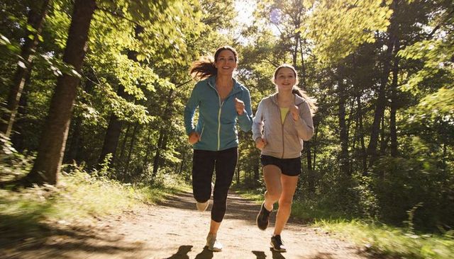 Mother and daughter jogging together on sunlit woodland trail, healthy outdoor fitness