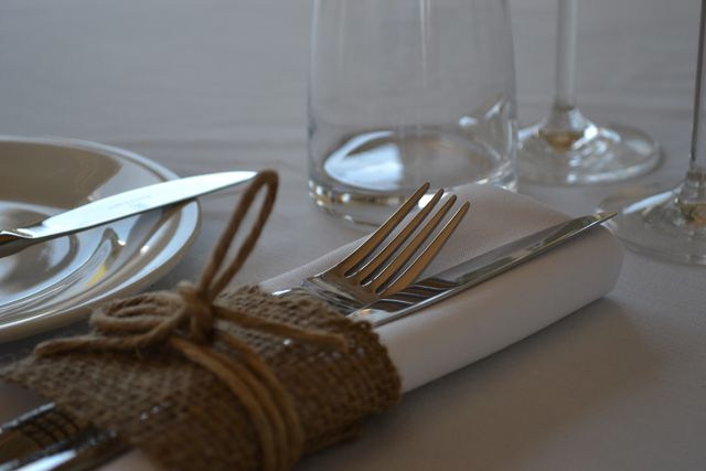 This photo showcases an elegant table setting with cutlery wrapped in a burlap cloth, placed on a white napkin. The presence of a clear drink ware and a neatly presented plate enhance the refined dining ambiance. This image is perfect for use in restaurant menus, dining advertisements, event planning guides, or blogs about table decor and dining etiquette.