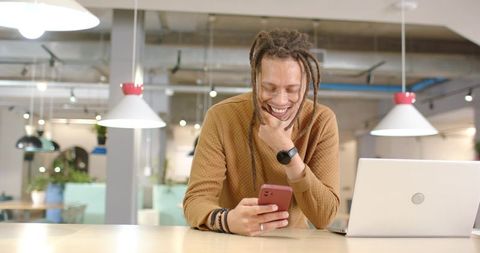 African American Man Leaning at Modern Counter Smiling Checking Smartphone with Laptop