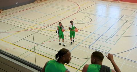Two basketball players holding balls walking across indoor hardwood court from balcony