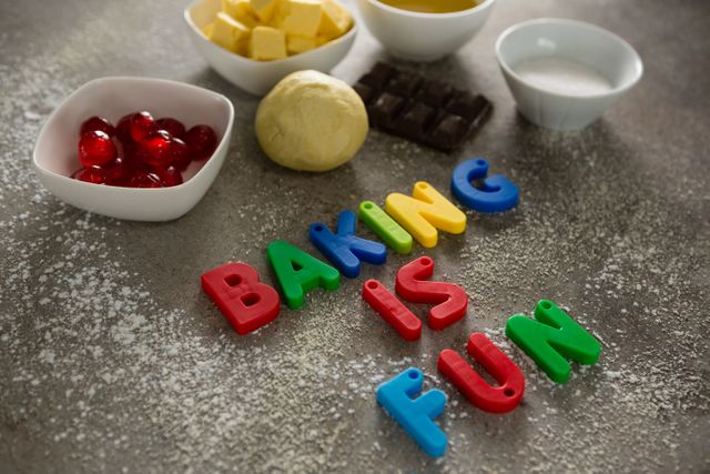 Various baking ingredients such as dough, butter, chocolate, sugar, and cherries are arranged on a table with colorful alphabet letters spelling 'Baking is Fun'. This image is perfect for illustrating baking activities, cooking classes, food blogs, and hobby-related content.