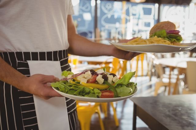 Mid section of waiter serving salad and French fries with burger in cafe