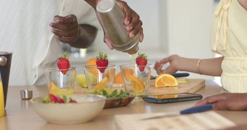 African american friends pouring citrus cocktails at kitchen counter with fresh fruit