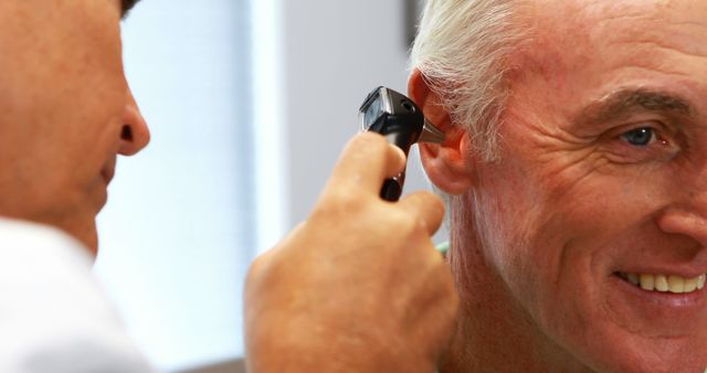 Senior man smiling while doctor uses otoscope to examine ear during routine medical checkup. Useful for illustrating medical examinations, healthcare professionals, ear care services, hearing tests, and senior patient care scenes.