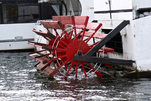Image depicts a close-up view of a red paddle wheel of a riverboat in contact with the water. This can be used for themes related to water transportation, mechanical features of boats, historical travel, and ferry services. Useful for illustrating articles, blog posts, and educational materials about boat mechanics, water-based transport, and scenic river cruises.