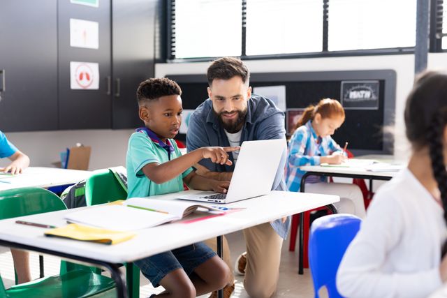 This image depicts a smiling teacher assisting a schoolboy with a laptop in an elementary school classroom. The scene highlights the use of technology in education and the supportive role of teachers. Ideal for use in educational materials, school websites, and articles about modern teaching methods, student engagement, and classroom technology.