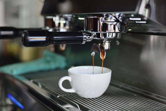 Picture showcases a professional espresso machine brewing fresh coffee into a white cup in a modern cafe setting. Ideal for use in articles, blog posts, or marketing materials related to coffee shops, barista training, or gourmet coffee experiences. Could also enhance brochures, websites, or social media posts highlighting café ambiance, coffee culture, or professional coffee equipment.