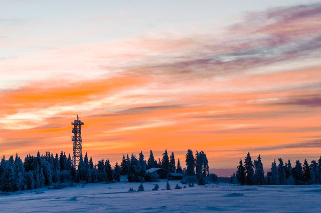 A beautiful winter sunset casts warm colors over a snowy landscape featuring a communication tower and snow-covered trees. This scene evokes tranquility and is ideal for use in winter travel content, nature articles, and relaxing background images.