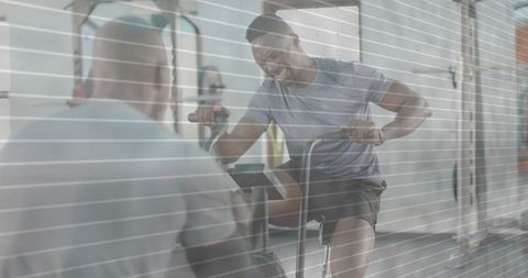 Male athlete pedaling stationary bike during personal training session through gym glass