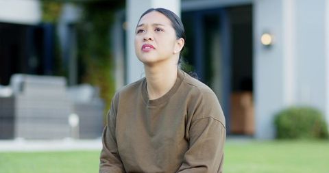 Thoughtful Asian Woman Sitting on Lawn by Modern Home