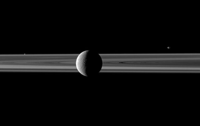 A pair of small moons join Saturn second largest moon in this NASA Cassini spacecraft image spotlighting Rhea in front of the rings. Janus is seen beyond the rings on the right and Prometheus is visible between the main rings and thin F ring on left.
