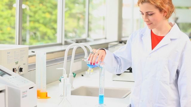 Young scientist in white lab coat carefully pouring blue liquid from one vessel to another in a bright, well-equipped laboratory. Perfect for illustrating concepts in scientific research, education, and innovation, or promoting chemistry and laboratory equipment brands.