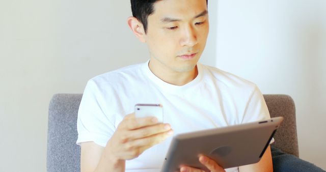 Young man sitting on couch, multitasking with smartphone and tablet. Ideal for concepts related to technology, communication, home office, and digital lifestyle.