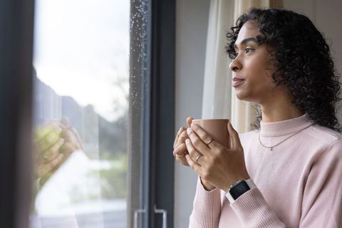 Non-binary person holding mug and gazing out window on rainy morning, quiet reflection