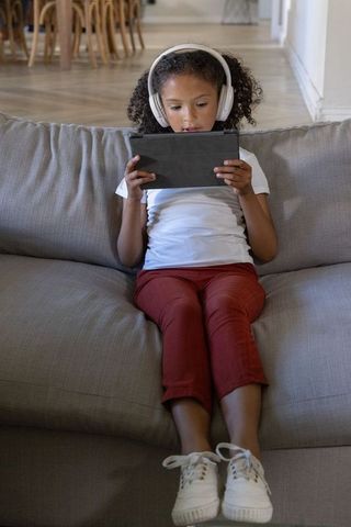 African american girl wearing headphones using tablet on sofa at home