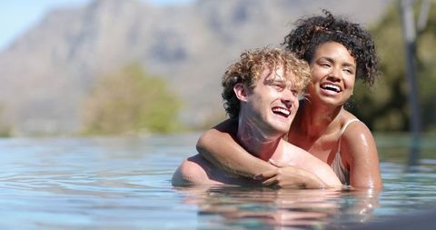 Sunlit romantic couple laughing and embracing in infinity pool with mountain view
