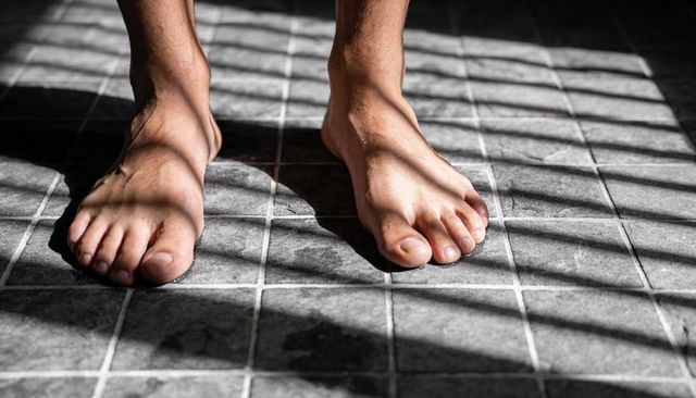Bare adult feet standing on gray square tiles with dramatic blind shadows and visible veins