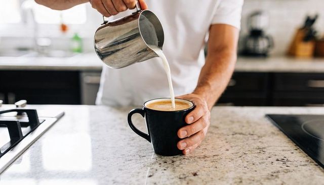 Pouring milk into latte mug on modern kitchen countertop, hands crafting coffee foam