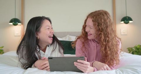 Laughing diverse friends sharing tablet on bed, cozy minimalist bedroom moment with plants
