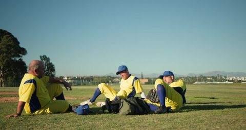 Male baseball teammates resting and talking on outfield grass wearing yellow blue uniforms