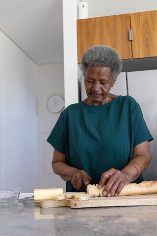 Senior woman slicing baguette on wooden board in bright kitchen preparing buttered bread