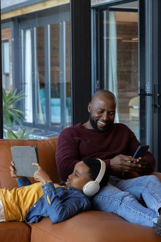 African american father and son relaxing on leather sofa using phone and tablet at home