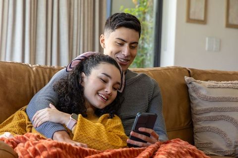 Diverse couple cuddling on leather couch sharing smartphone during cozy relaxed afternoon