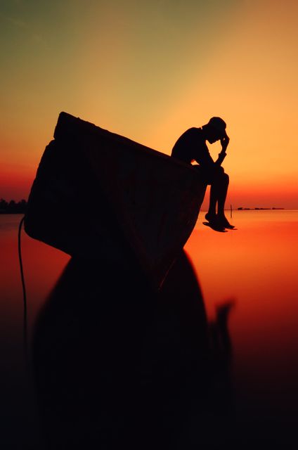 Silhouette of a man sitting on a boat during sunset, creating a tranquil and peaceful scene with a vivid orange and red sky in the background. This can be used for travel blogs, meditation guides, or nature websites.