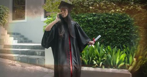 Graduate in cap and gown talking on phone while holding diploma on campus steps