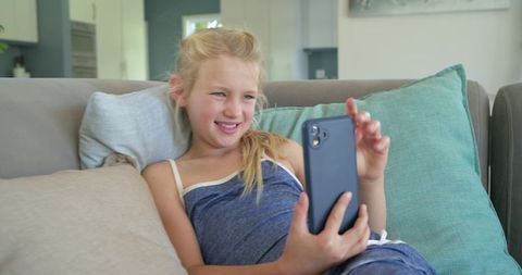 Preteen girl relaxing on couch using smartphone, smiling in cozy modern living room