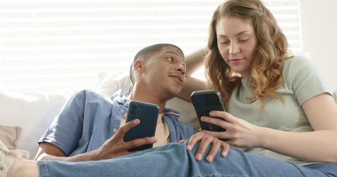 Mixed-race couple relaxing on couch, sharing smartphones and enjoying cozy at-home moment