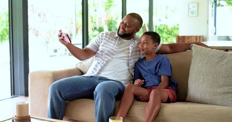 African american father taking selfie with son on cozy living room sofa, smiling