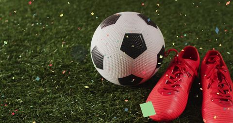 Soccer ball and red shoes with falling confetti on grass field