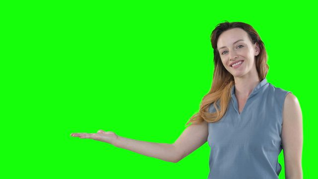 This image features a happy Caucasian woman with long brown hair, dressed in a grey blouse, confidently presenting on a green background. She has her arm raised, and her open palm beckons as though she is introducing or showcasing virtual content. Perfect for use in presentations, advertising concepts of technology or interactive screens, or projects needing customizable areas for texts and graphics overlaying the green background.