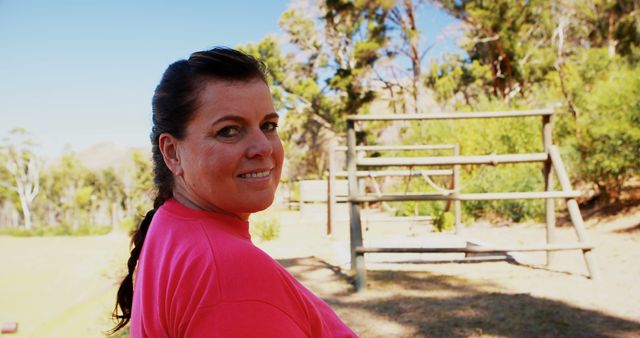 Middle-aged woman smiling at camera while standing outside near an obstacle course. Ideal for content related to outdoor fitness, health initiatives, active lifestyle, and motivational materials. Shows confident demeanor and natural outdoor setting.