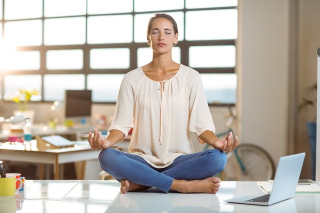 This image depicts a businesswoman practicing meditation in an office to relieve stress and promote mindfulness. Ideal for websites and articles on corporate wellness, mental health, stress management, and work-life balance. Useful for illustrating professional settings that integrate relaxation techniques into the workplace.