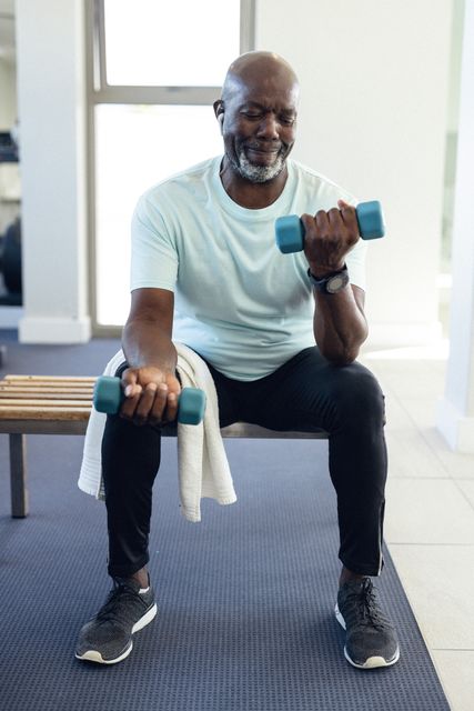 African American man sitting on a bench, lifting dumbbells with a smile. Ideal for promoting home fitness, healthy lifestyle, senior fitness programs, and wellness campaigns.