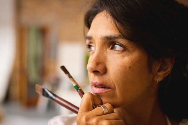Biracial female artist holding paintbrushes, looking thoughtful in her studio. Ideal for use in articles or advertisements about creativity, artistic inspiration, and the life of an artist. Suitable for promoting art supplies, creative workshops, or artist profiles.