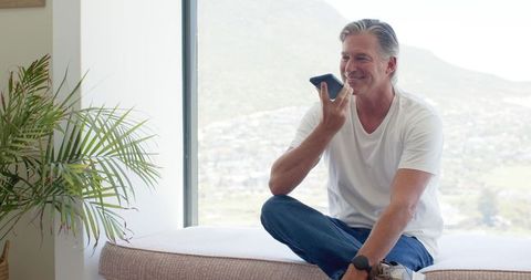 Mature man sitting by window speaking on smartphone smiling relaxed modern home interior