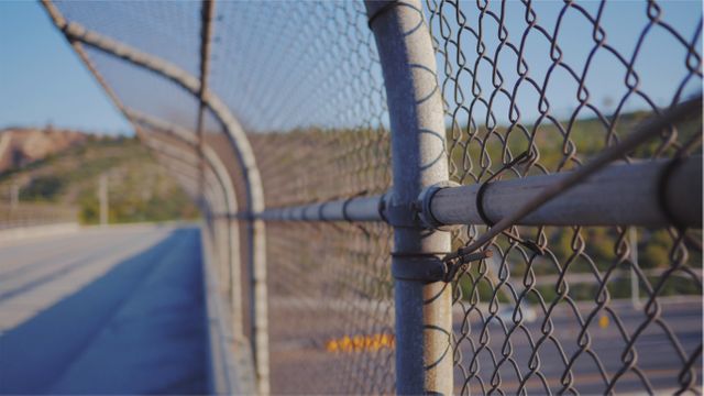 Showing a rusty chain link fence with a blurry mountainous background and clear blue sky, highlighting themes of security and rustic outdoor environment. This could be used in designs relating to outdoor activities, safety, or rustic landscapes.