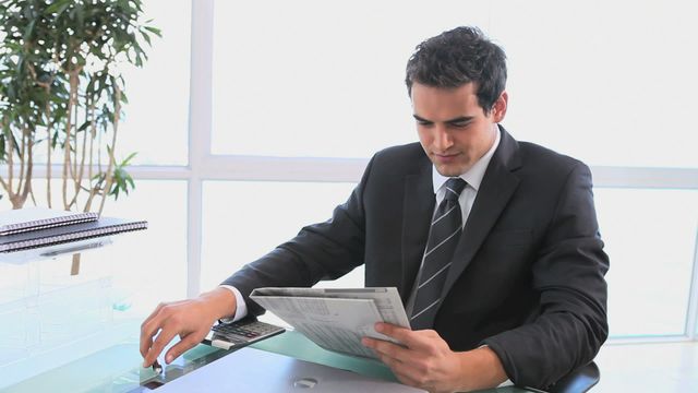 Businessman in a sharp suit engrossed in reading a newspaper in a bright, spacious office. Sunlight enters through large windows, illuminating the modern decor and elegant arrangements. Useful for content about corporate culture, office dynamics, professional lifestyle, or business news updates.