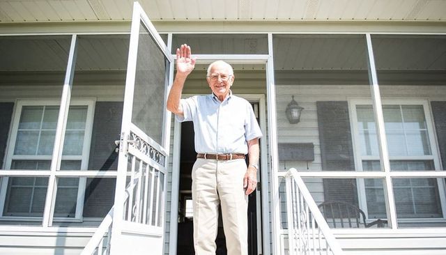 Smiling senior man waving from screened porch doorway on sunny day, home independence