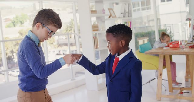 Two boys are shaking hands in a modern office. Both are dressed in formal attire, one in a blue sweater with tan pants and glasses, and the other in a navy suit with a red tie. They look like young professionals possibly role-playing or engaged in a business-related activity. This scene can be used for depicting themes of teamwork, youth in business, early education, professional growth, and future leaders. A girl can be seen in the background, possibly involved in another activity.