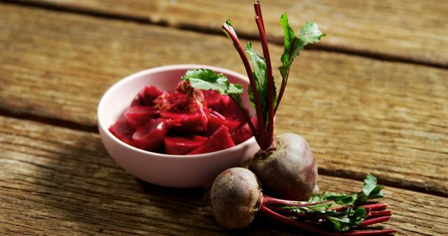 A bowl of chopped beets is placed on a wooden surface alongside whole beets with leafy greens attached. Beets are known for their deep red color and are often used in healthy cooking for their nutritional benefits.